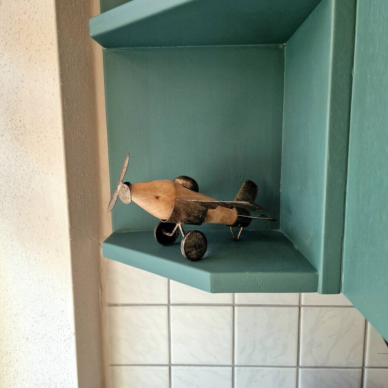 Un petit modèle d’avion en bois est posé sur une étagère de couleur turquoise dans une salle de bain dont les murs sont recouverts de carreaux blancs.
