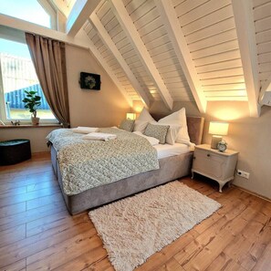 Une chambre avec un plafond en pente, un lit recouvert d’une couvre-lit à motifs, une table de chevet avec une lampe et une fenêtre avec des rideaux.