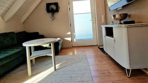 A cozy attic room with a high ceiling, a large window, a flat-screen TV, a green couch, a small table, and a white cabinet. - Select-Home 6 - Haus Altstadt (Neunburg vorm Wald)