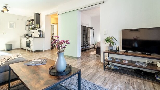 A modern living room with a wooden coffee table, a vase with flowers, a flat-screen TV, and a bookshelf. - Select-Home 2 -Netflix | parking lot | many extras (Bruck in der Oberpfalz)
