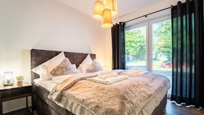A modern bedroom with a large bed, dark headboard, and a view of the outdoors through a window with curtains. - Select-Home 2 -Netflix | parking lot | many extras (Bruck in der Oberpfalz)