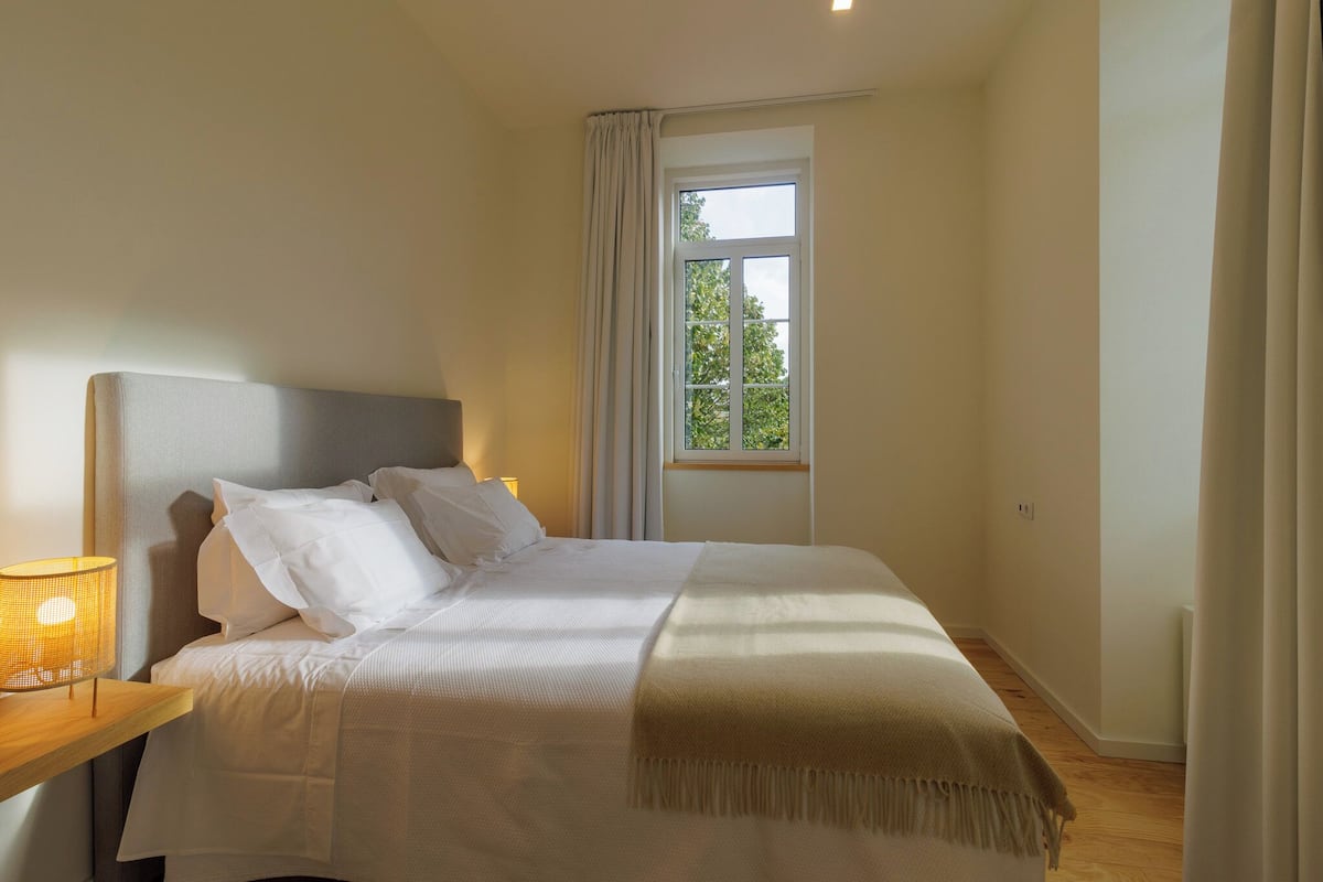 Een netjes opgemaakt bed met witte lakens en een plaids, een bedlampje en een raam met gordijnen.