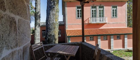 A covered outdoor seating area with a table and chairs, overlooking a building with a red-tiled roof and a balcony.