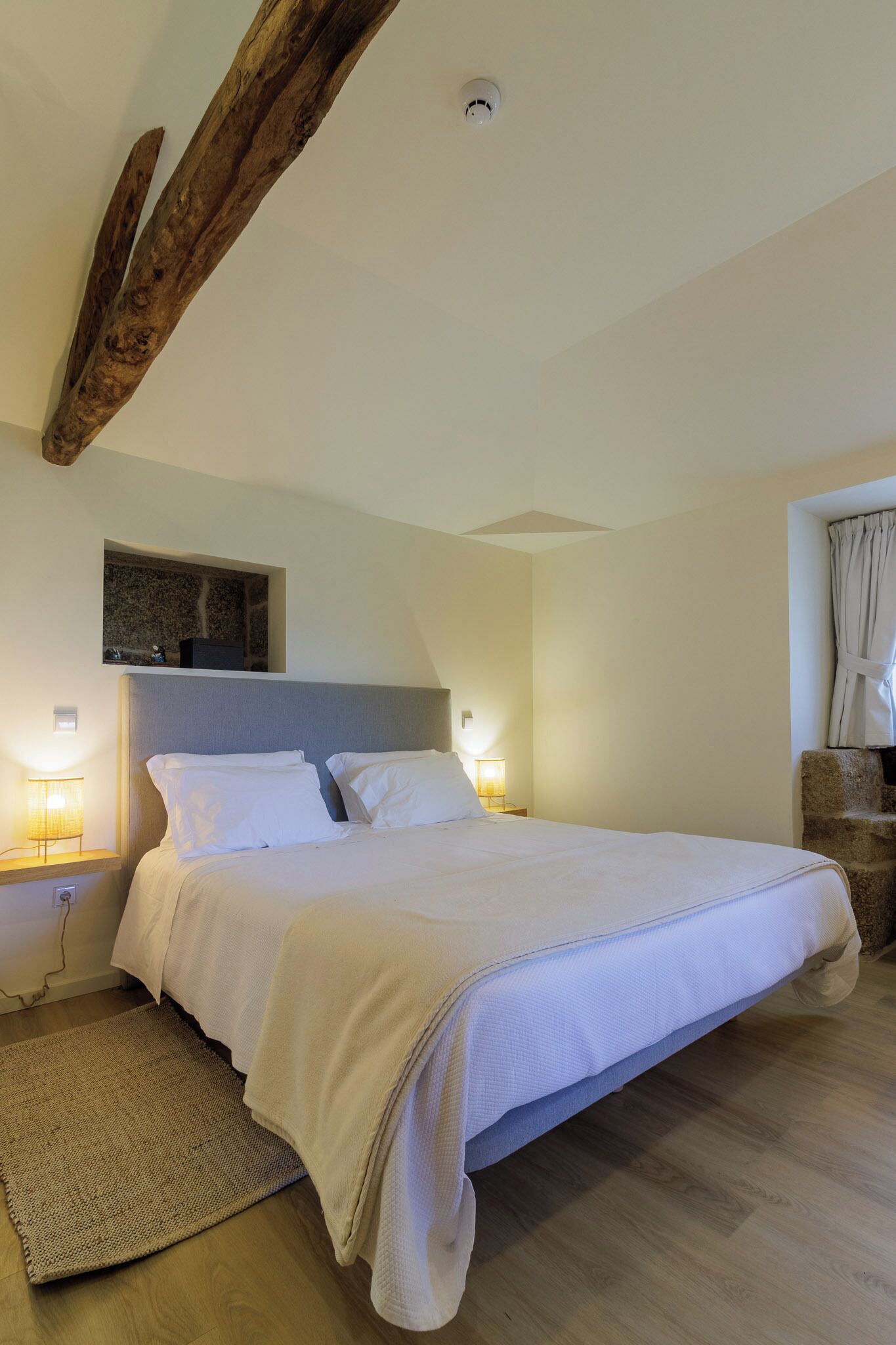 A bedroom with a large bed, wooden beams, a stone wall, and a window with curtains.