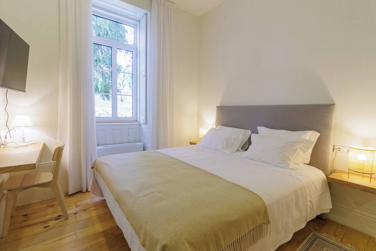 Een netjes opgemaakt slaapkamer met een bed, nachtkastje, stoel en een aan de muur bevestigde televisie.