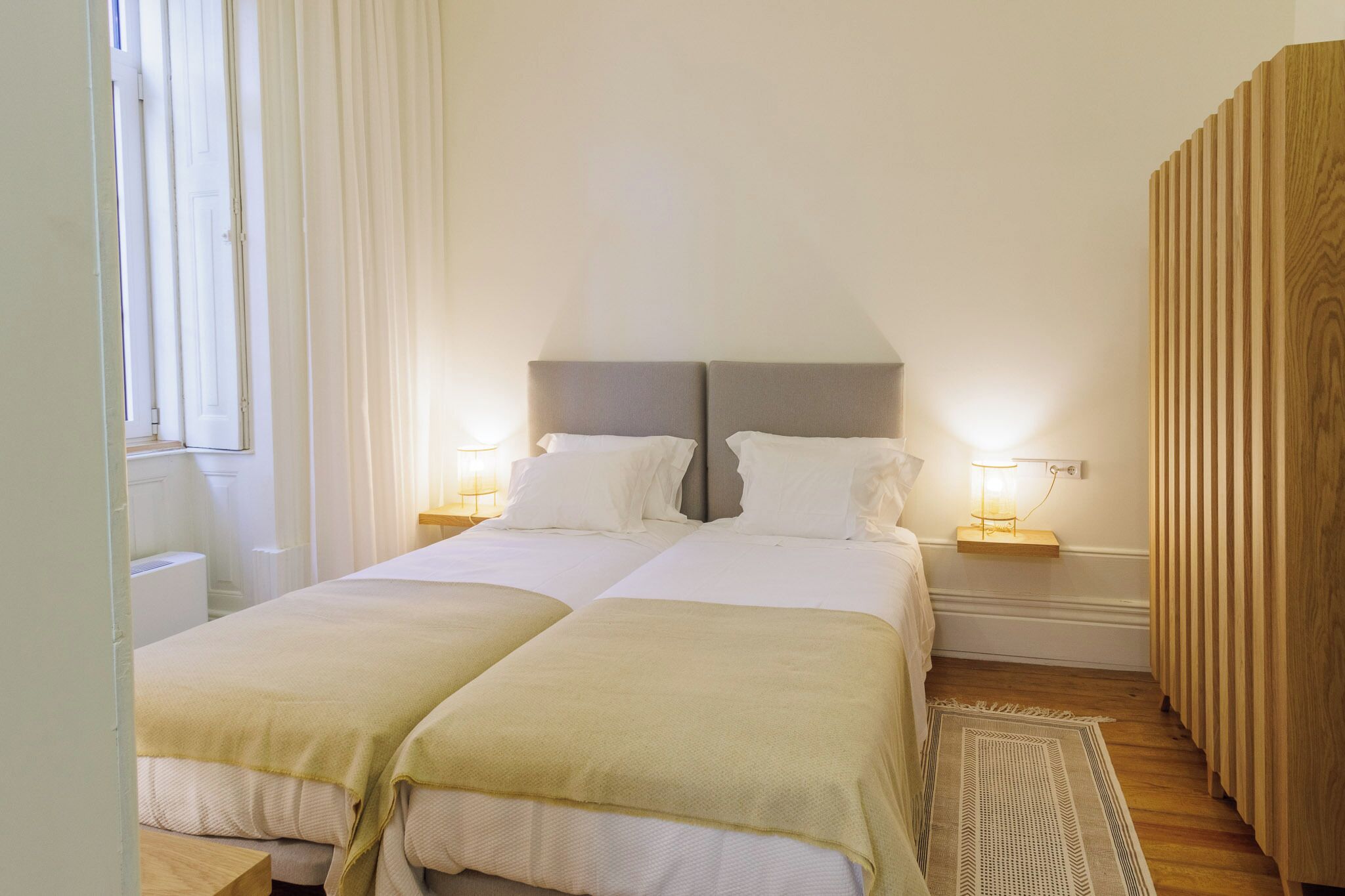 A double room with two beds, each with a bedside table and lamp, a wooden wardrobe, and a window with curtains.