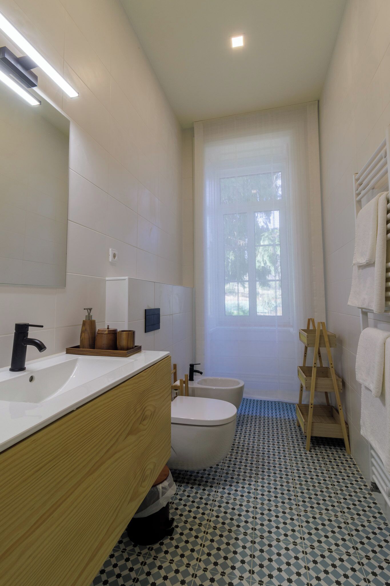 Un baño moderno con un lavamanos blanco, un grifo negro y un mueble de tocador de madera. Hay un inodoro sobre un suelo con diseño azul, y una ventana con cortinas blancas.
