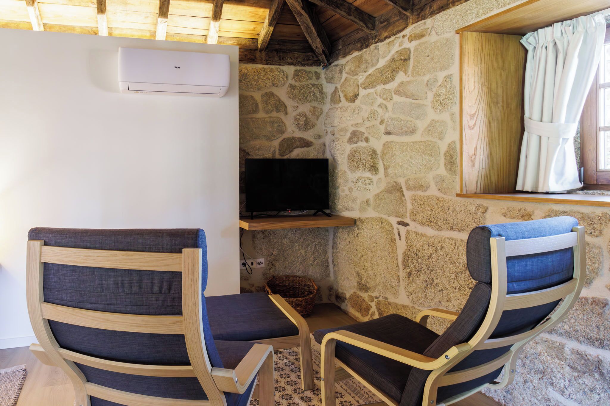 A cozy room with two wooden chairs with cushioned seats, a stone wall, a television on a wooden shelf, and a window with curtains.