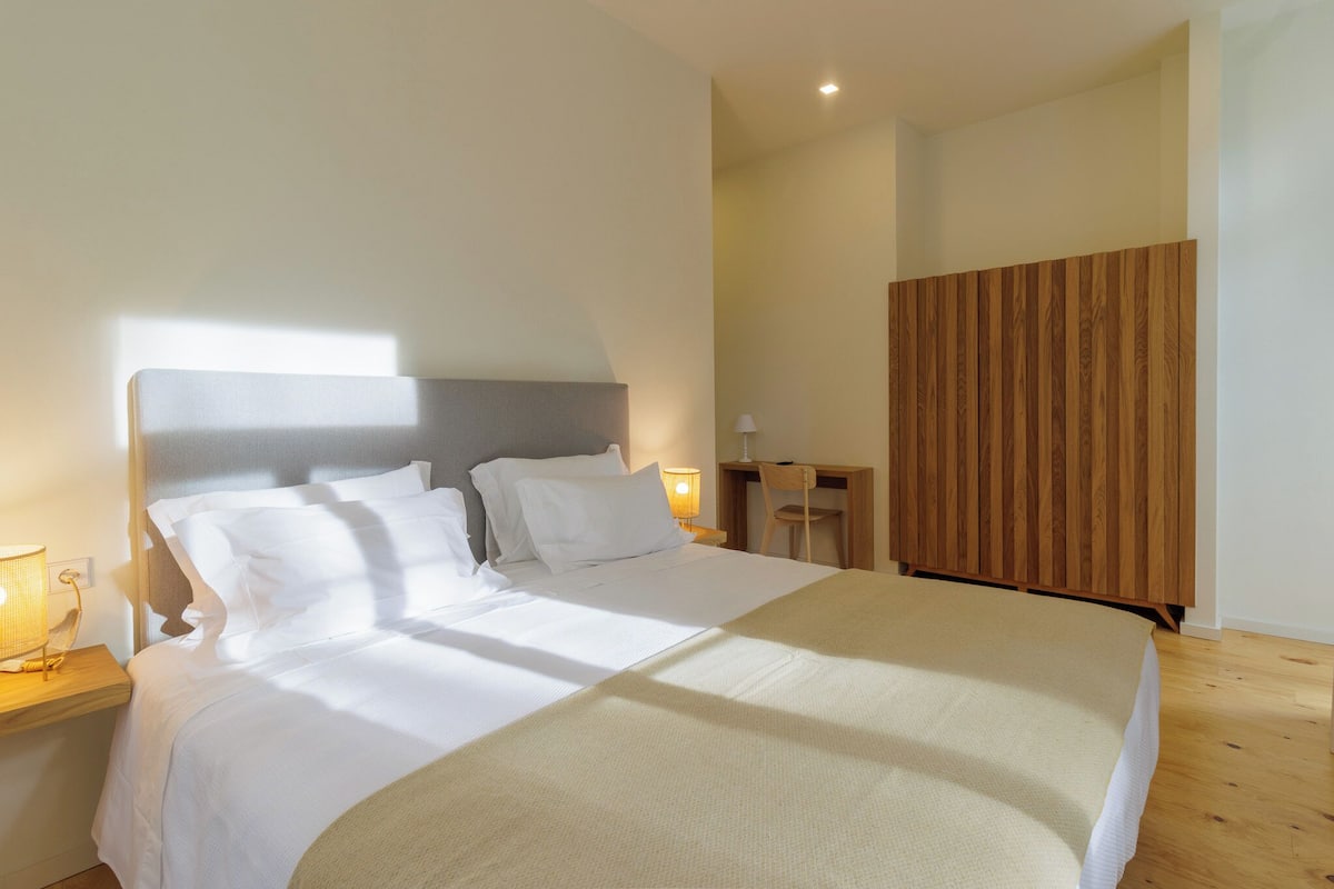 Een moderne hotelkamer met een groot bed, een houten hoofdbord, een nachtkastje met een lamp en een klein bureau met een stoel.