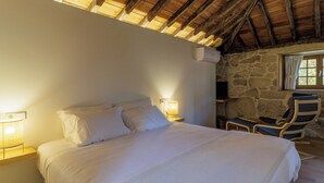 Een slaapkamer met een groot bed, twee bedlampjes, een stenen muur, een raam met gordijnen en een stoel met kussen.