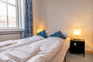 A double bed with white linens, a bedside table with a lamp, and a window with curtains.