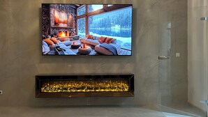Una sala de estar moderna con un televisor de pantalla plana grande montado en la pared, una chimenea debajo y un florero con rosas rojas sobre una mesa de vidrio.