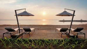 Terraza de madera con sillones de descanso y sombrillas, con vista a una masa de agua tranquila al atardecer.
