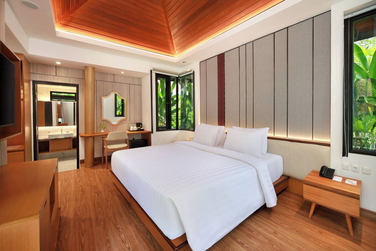 A modern hotel room with a large bed, wooden flooring, a desk, a chair, a vanity with a mirror, and a window with a view of greenery.