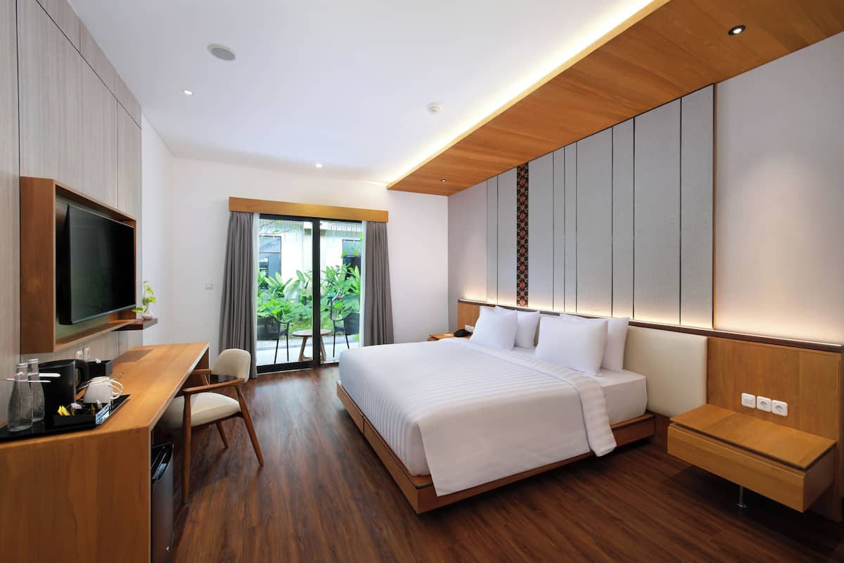 A modern hotel room with a large bed, wooden flooring, a desk with a flat-screen TV, and a sliding glass door leading to a balcony.