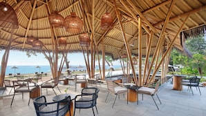 Zona de estar al aire libre con estructuras de bambú, mesas y sillas, con vistas a una playa.