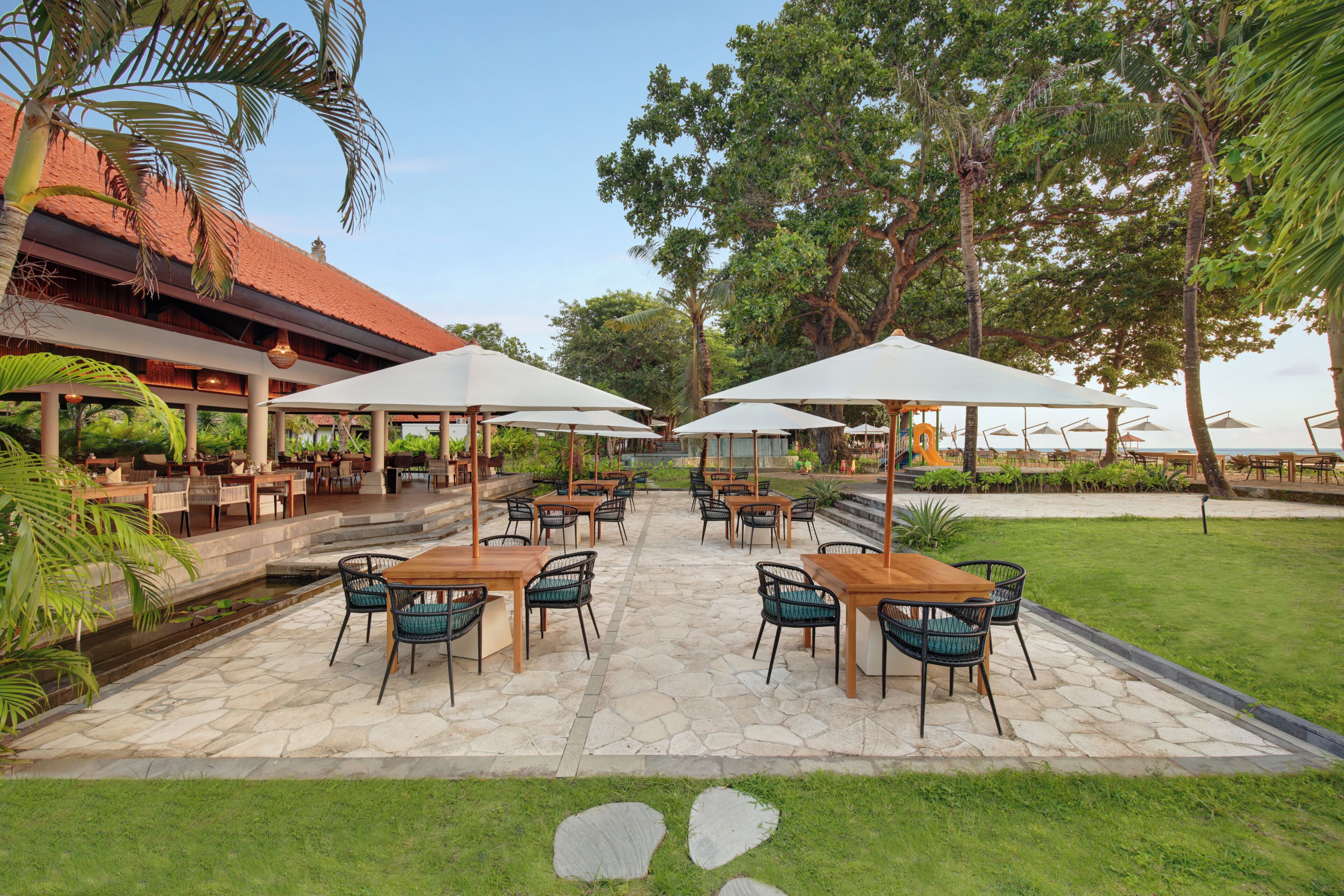 An outdoor patio with stone flooring, wooden tables, and green chairs, surrounded by lush greenery and trees.