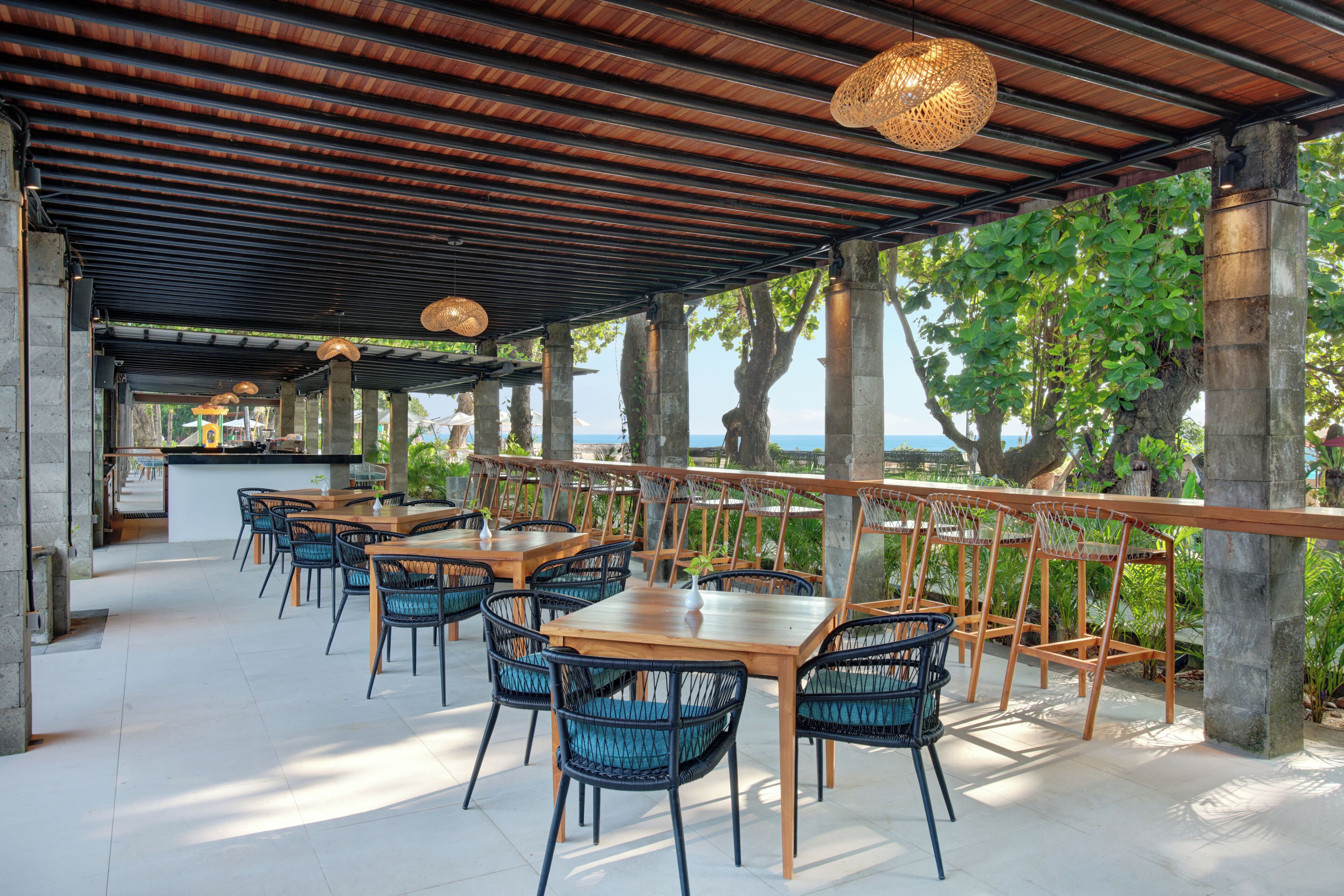 An outdoor dining area with wooden tables and chairs, a covered roof, and a view of the sea.