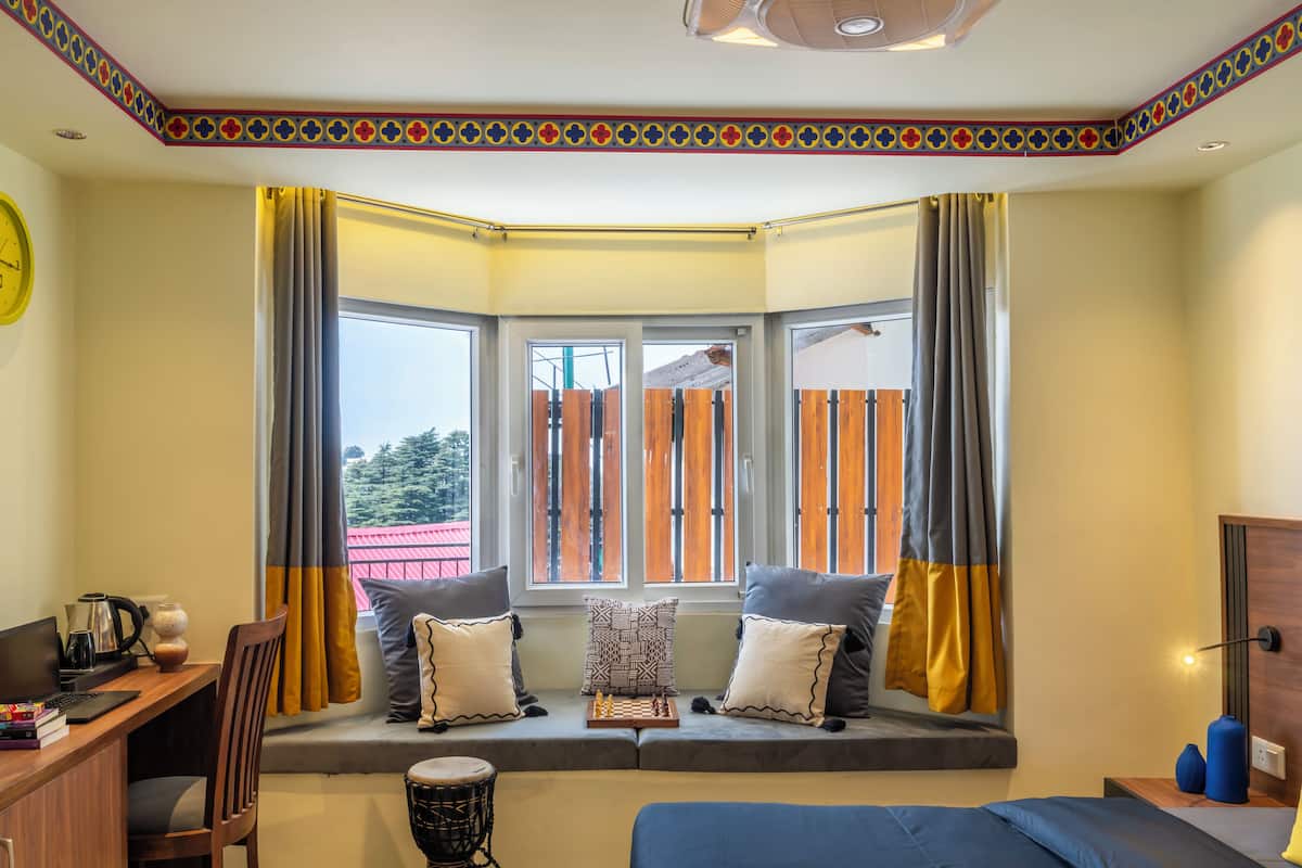 A room with a bay window, a desk with a laptop, a chair, a bed, and a small table with a lamp.