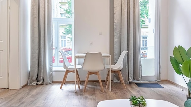 A modern room with a dining table and chairs, large windows with curtains, a potted plant, and a view of the street outside. - Lovely Apartment Steps from Schonbrunn (Vienna)