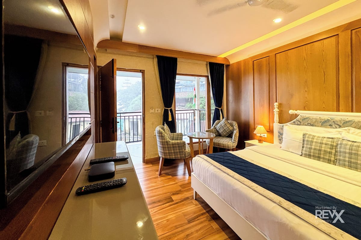 Ein Hotelzimmer mit Bett, Schreibtisch inklusive Fernbedienungen, einem Sessel, einem kleinen Tisch und einem Balkon mit Vorhängen.