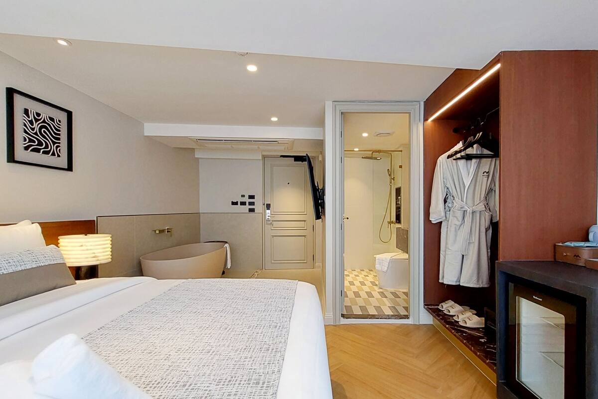 A hotel room with a bed, bedside table, lamp, robe, and a shower area.