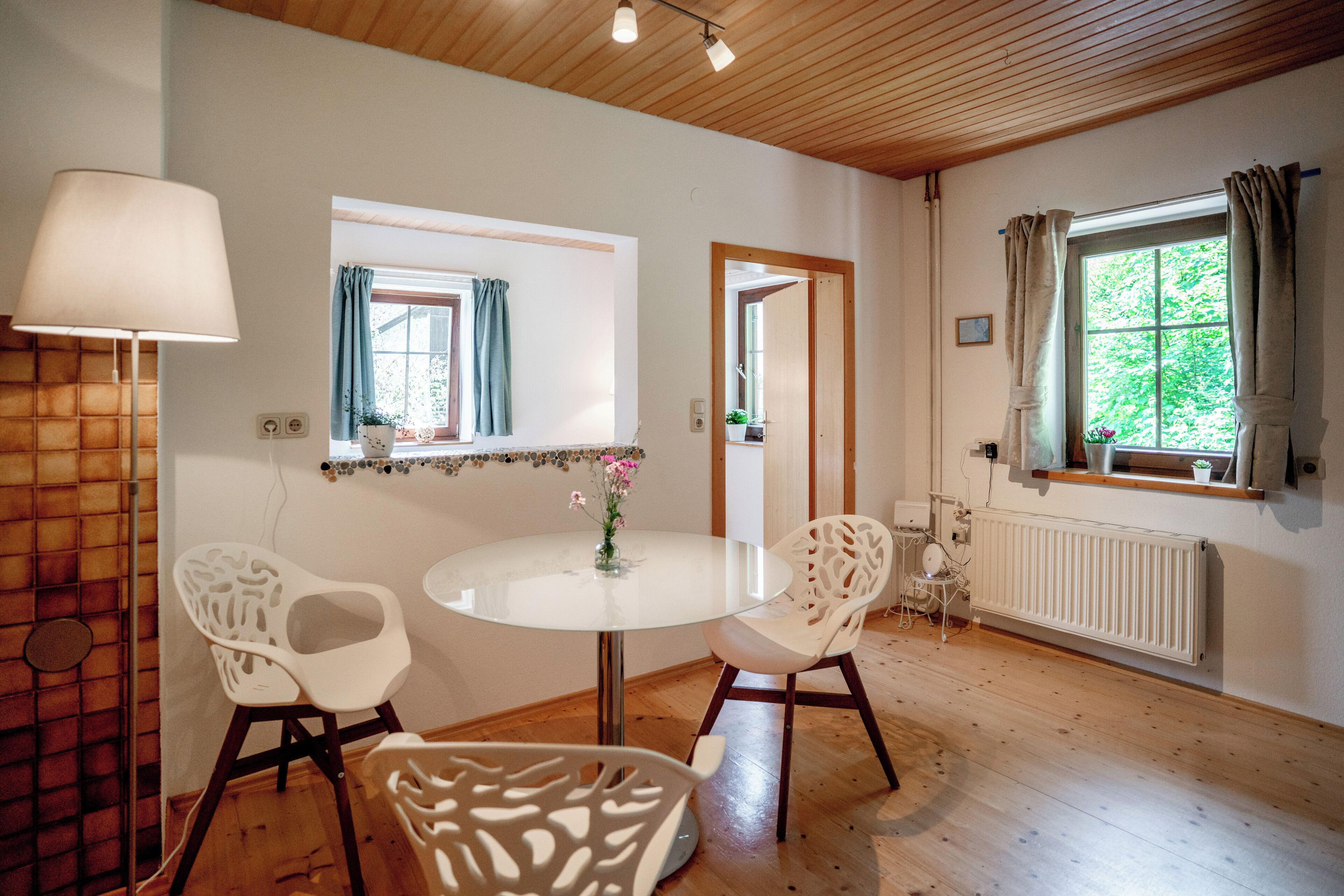 A room with a round glass table, two white chairs, a lamp, a window with curtains, a radiator, and a wooden ceiling.