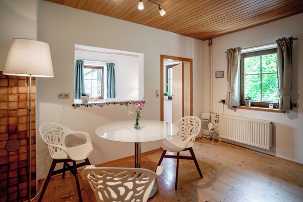 A room with a round glass table, two white chairs, a lamp, a window with curtains, a radiator, and a wooden ceiling.