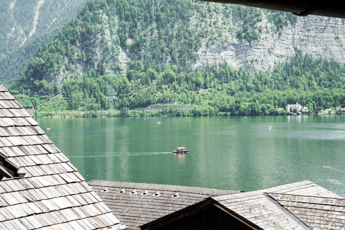 A view of a lake with a boat, surrounded by forested mountains and a building with a sloped roof.