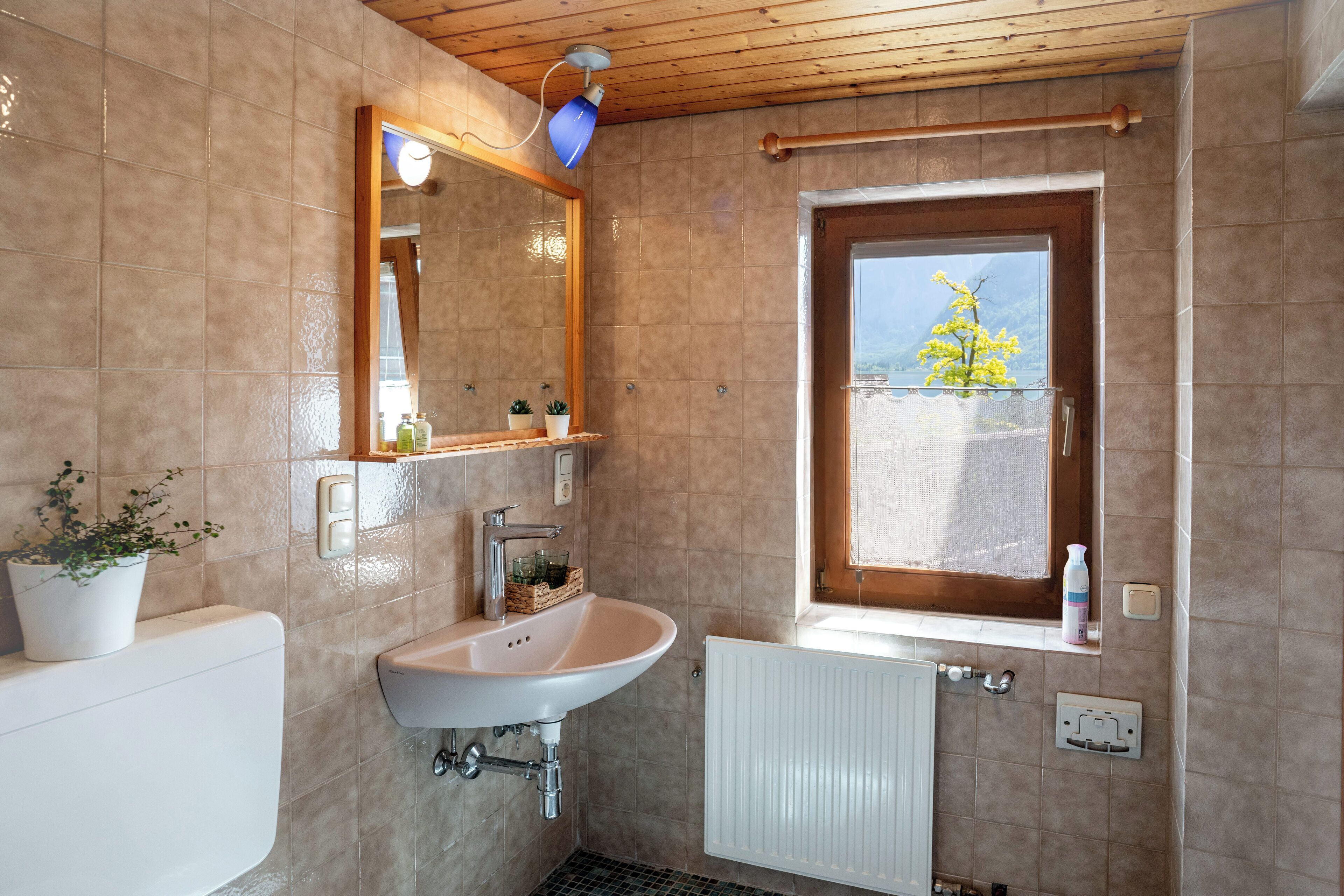 Une salle de bain avec un lavabo blanc, un miroir, une fenêtre donnant sur un immeuble et un arbre, et un plafond en bois.