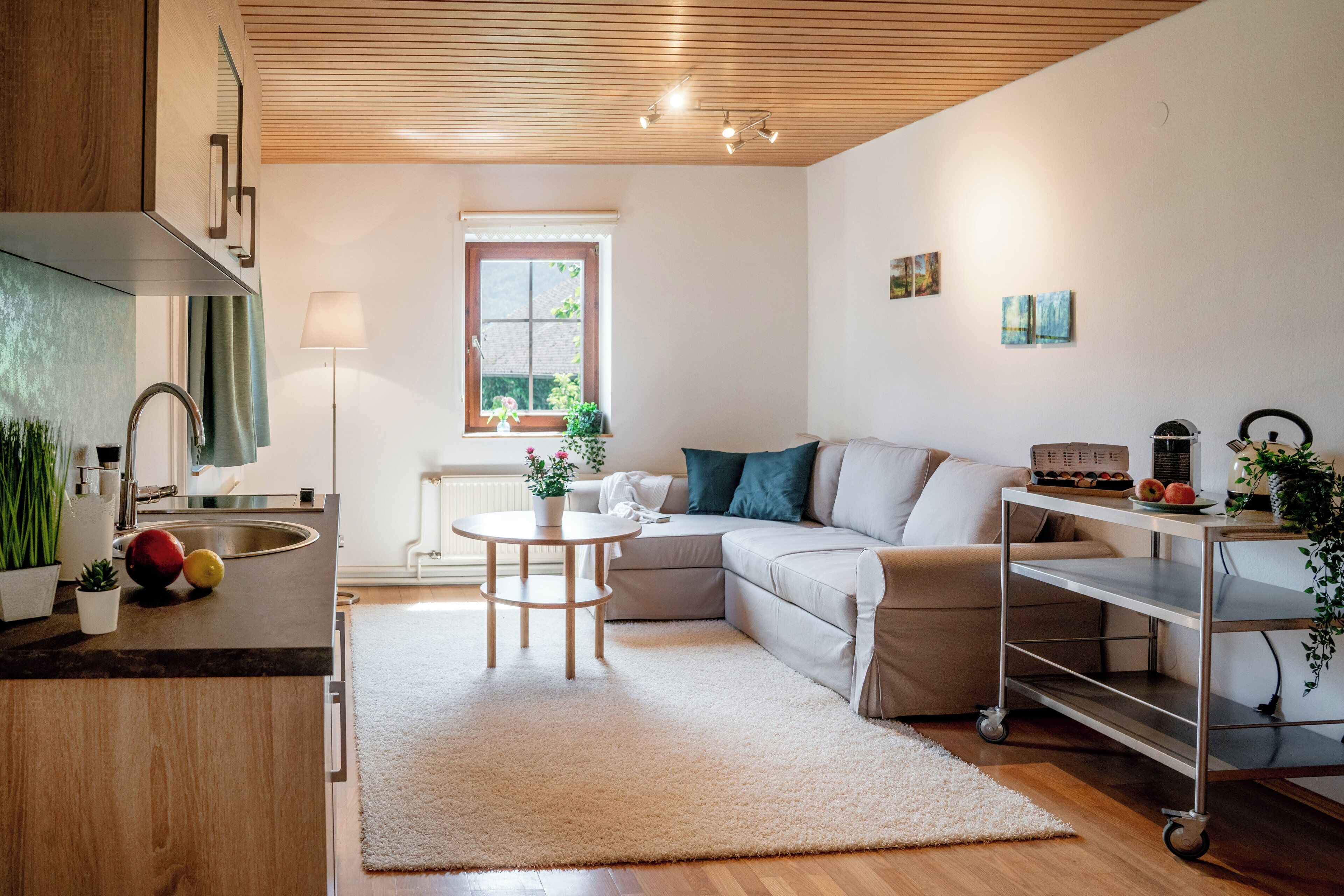 A modern kitchen with a wooden ceiling, a dining area with a white table and chairs, a sofa with teal cushions, and a cart with fruits and a plant.