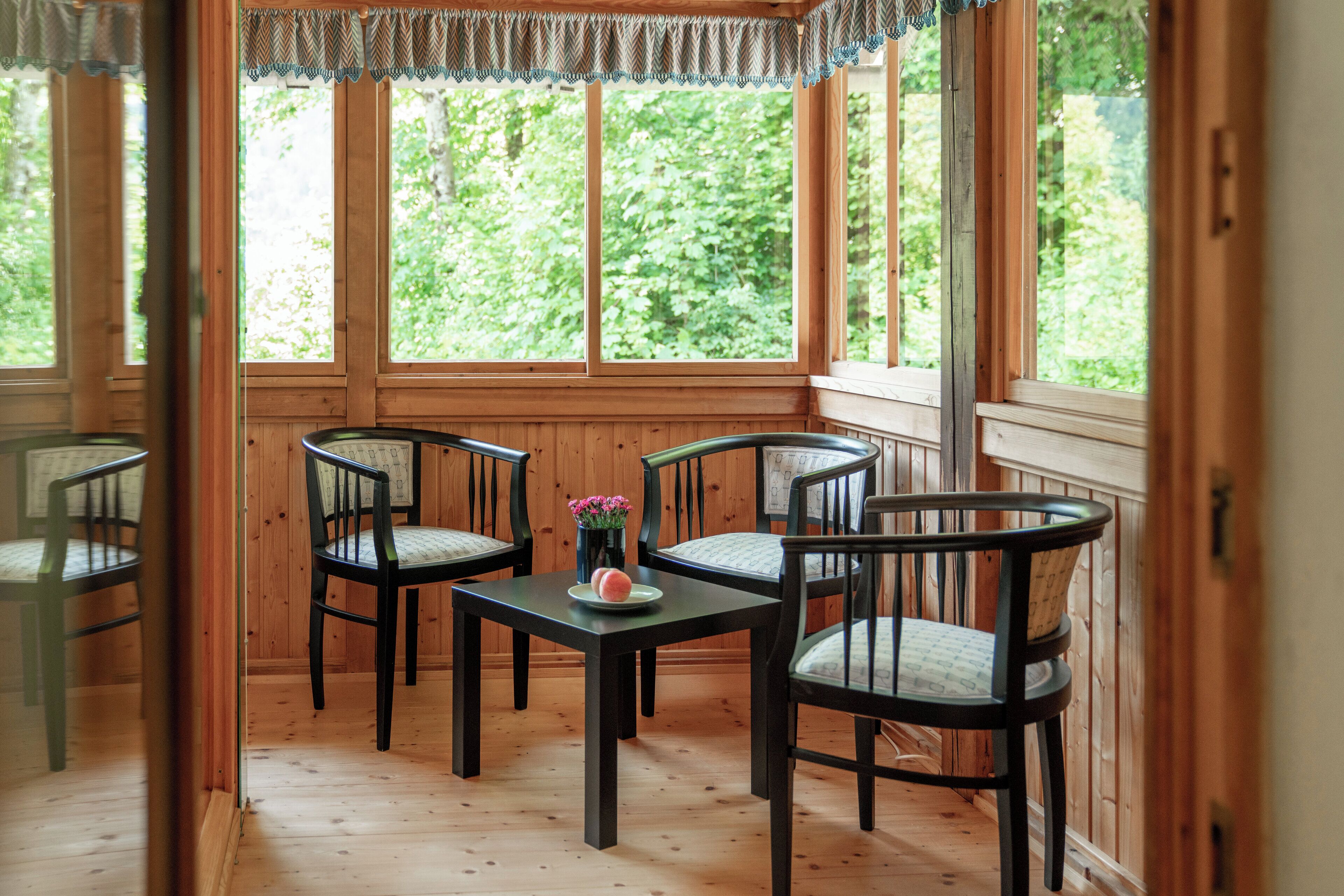 Una habitación con paneles de madera, una mesa y cuatro sillas, ventanas amplias y vistas al verdor exterior.