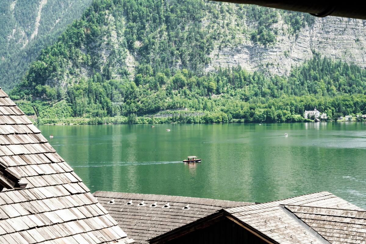A view of a lake with a boat, surrounded by forested mountains and a building with a sloped roof.