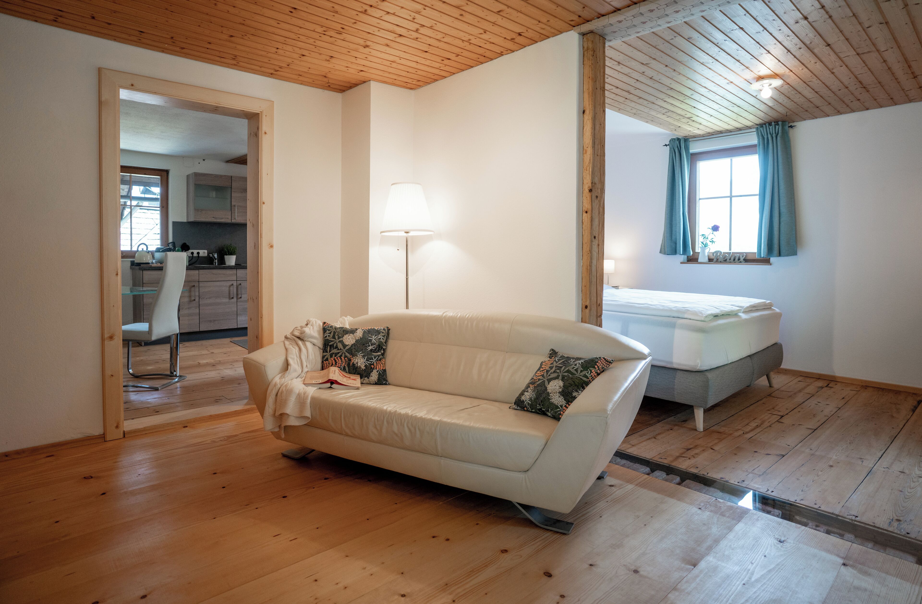 A room with a wooden ceiling, a white sofa with patterned cushions, a bedside table with a lamp, and a window with curtains.