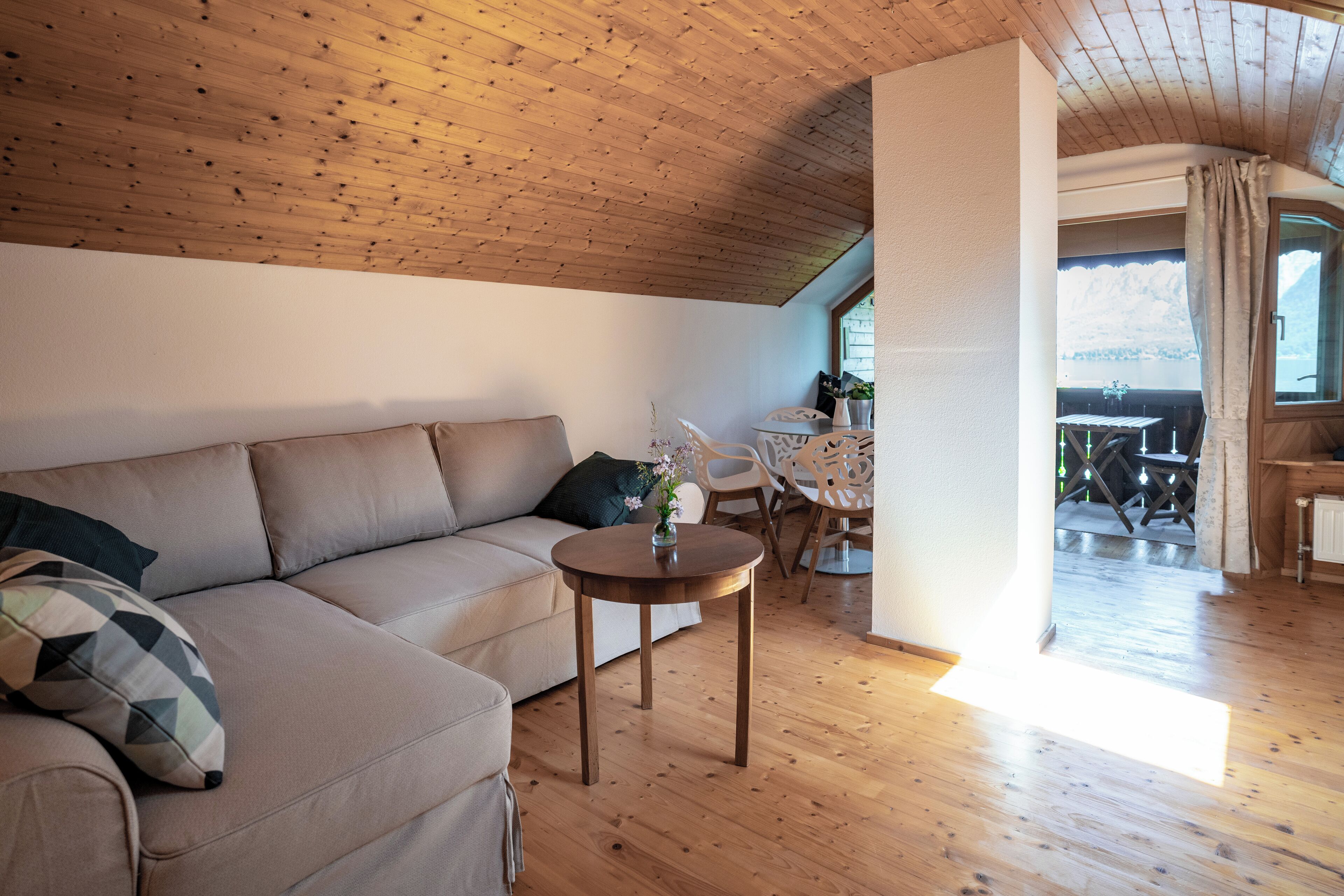 A cozy living room with a wooden ceiling, a beige sofa, a small round table with a vase, and a view of a balcony with outdoor furniture.