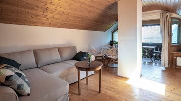 A cozy living room with a wooden ceiling, a beige sofa, a small round table with a vase, and a view of a balcony with outdoor furniture.