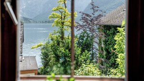A view through a window of a lake with mountains in the background, a wooden deck, and a house with a sloped roof. - Lakeview Apartments (Hallstatt)