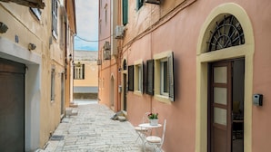 Une ruelle étroite pavée, bordée de bâtiments des deux côtés, avec des fenêtres et des portes en arc de cercle, et un petit espace extérieur aménagé avec une table et des chaises.
