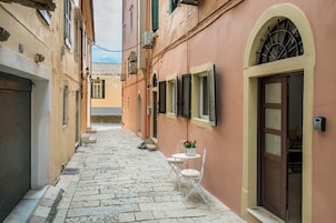 Una stretta strada acciottolata fiancheggiata da edifici con finestre e porte ad arco, e una piccola zona con tavoli e sedie.