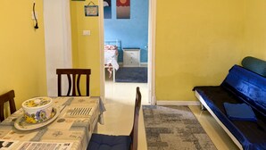 A dining area with a table set for two, a blue sofa, and a door leading to a bedroom with a bed and a nightstand. - Paint House (Imperia)