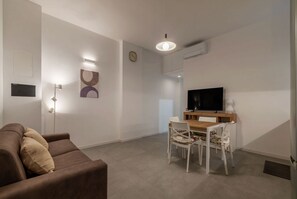 A modern living room with a brown sofa, a dining table with chairs, a flat-screen TV, and a ceiling light.