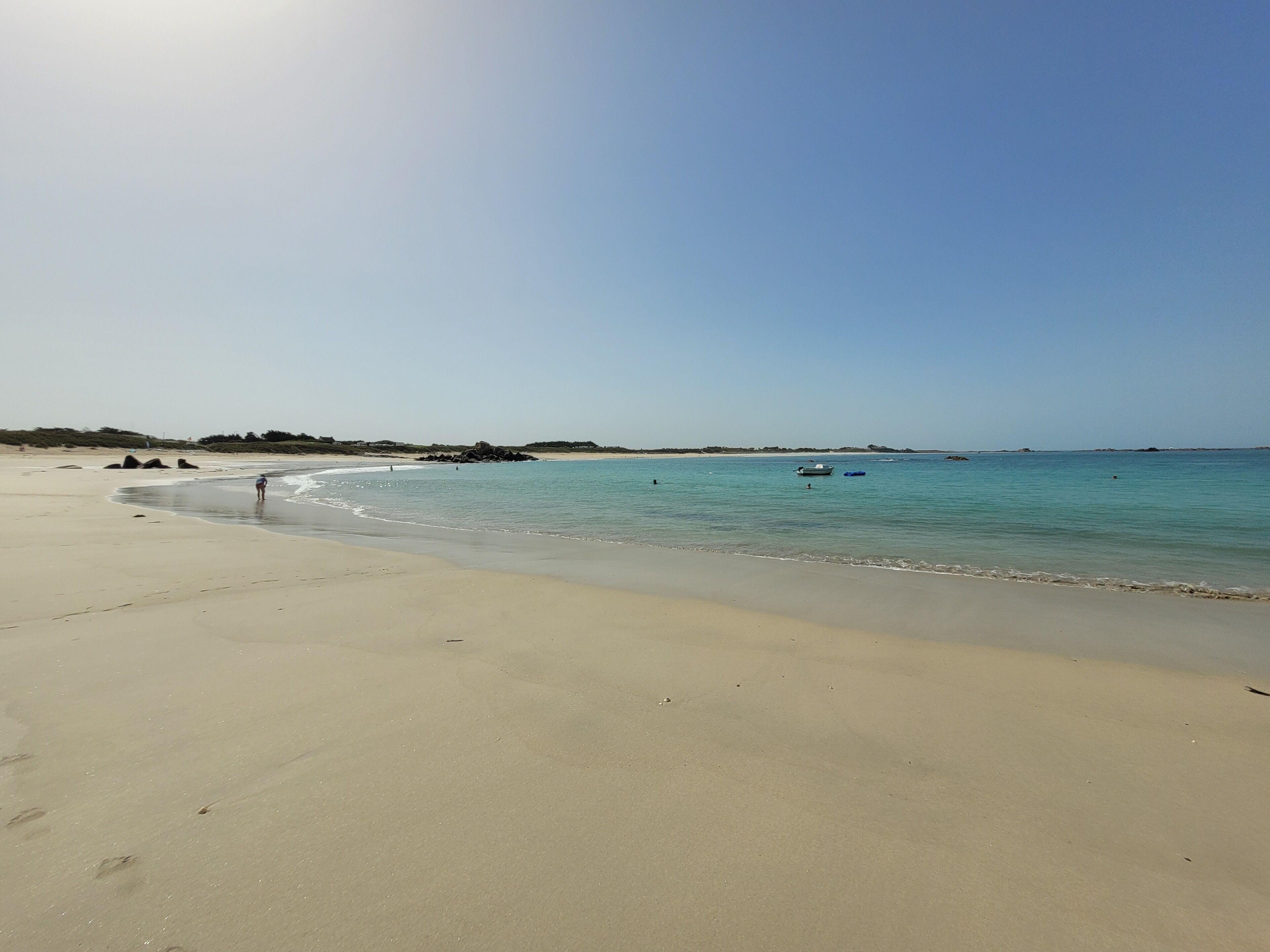 Una playa arenosa con aguas tranquilas, algunas embarcaciones y formaciones rocosas a lo lejos.