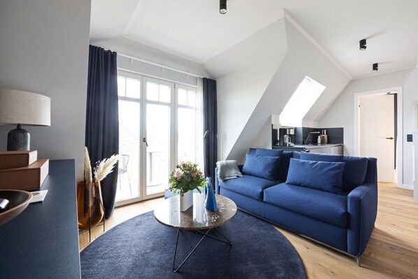 A modern living room with a blue sofa, a coffee table, and a view of the kitchen through a glass door. - Dünenloft, House Kliff Whg. 12 - Dünenlofts (Sylt)