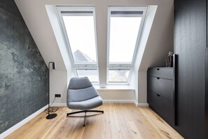 Een moderne kamer met een schuin dak, twee dakramen, een grijze bureaustoel, een zwarte vloerlamp en een donker houten dressoir.