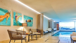 Un espace piscine intérieur moderne, doté de chaises longues, d’une table avec des fruits et d’œuvres d’art accrochées aux murs.