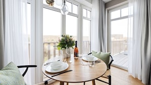 Un coin repas avec une table ronde en bois, des assiettes blanches, des couverts et une bouteille de vin. Il y a deux chaises avec des coussins, et un vase rempli de fleurs sur la table.