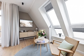 A modern loft bedroom with a sloped ceiling, large skylight windows, a wooden floor, a round table with a vase of flowers, a wicker chair, and a flat-screen TV on the wall. - Dünenloft, Haus Kliff, Whg. 9 - Dünenlofts (Sylt)
