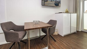 Una habitación moderna con una mesa de madera, dos sillas grises, un armario blanco y un televisor en la pared que muestra una escena de playa.