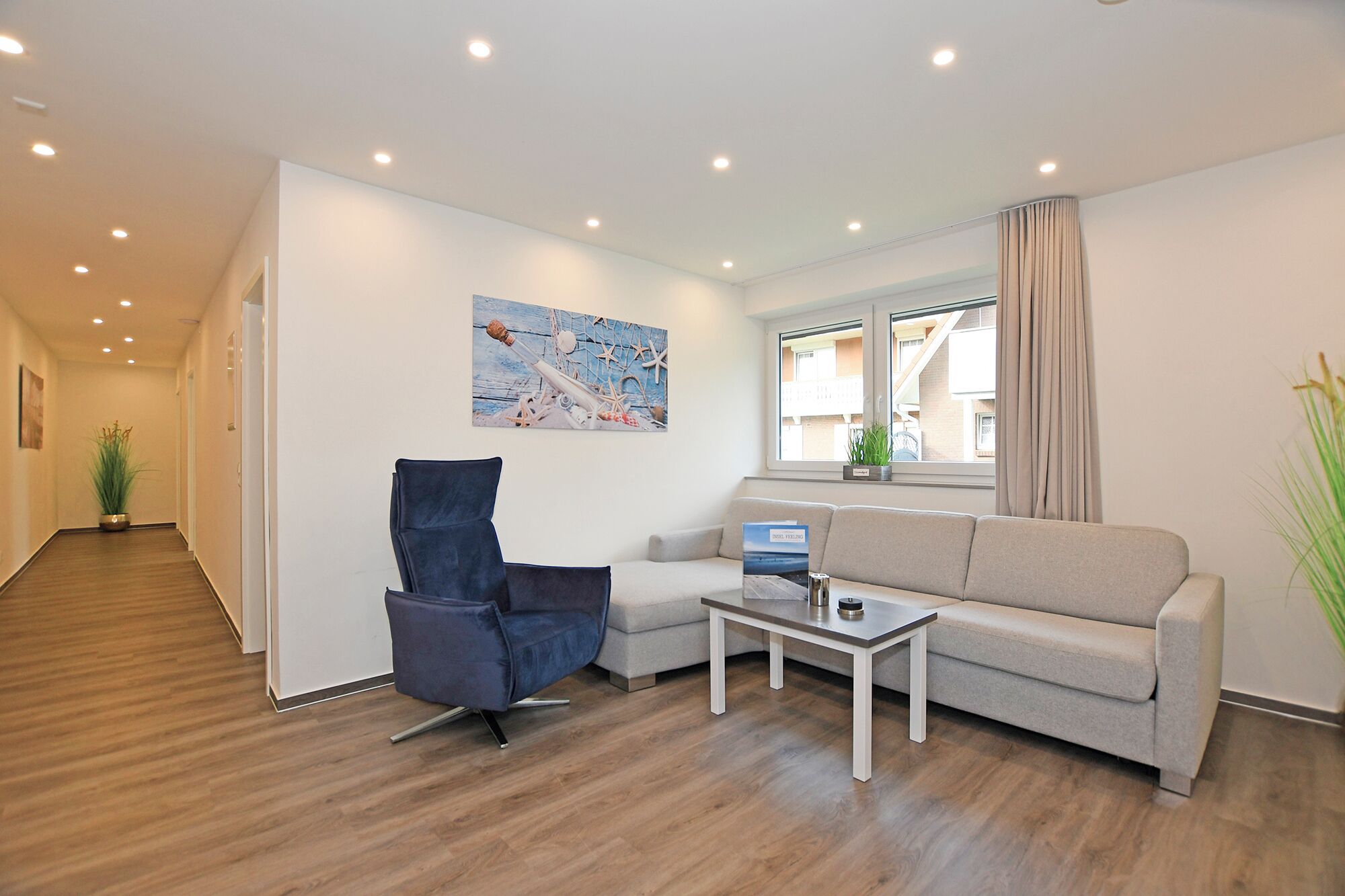 Una sala moderna con un sofá gris, un sillón azul y una mesita de centro. Hay un cuadro en la pared y una ventana con cortinas.