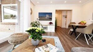 A modern living room with a large window, a dining area with a table and chairs, and a television mounted on the wall. - Island Feeling Apartment 2 (Büsum)